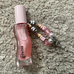 Può includere: Una bottiglia di vetro trasparente con un liquido rosa al suo interno. La bottiglia è etichettata "GISOU Honey Infused Lip Oil" e ha un ciondolo con perline rosa e bianche.