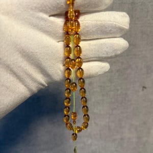 May include: A strand of amber-colored prayer beads. The beads are oval-shaped and strung on a green cord. The beads are a translucent, golden-brown color. The tassel at the end of the strand is also amber-colored.