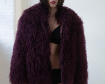 May include: A deep purple, shaggy faux fur coat. The coat is open, revealing a black bra and black bottoms. The model is standing in front of a white wall and a white brick wall.