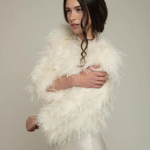 Könnte beinhalten: Cremefarbene Feder-Bolerojacke. Die Jacke hat einen runden Ausschnitt und Dreiviertelärmel. Die weiche, flauschige Textur der Federn erzeugt einen luxuriösen Look. Der Bolero wird über einem weißen Satinkleid getragen.