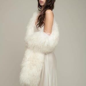 Pode incluir: Uma mulher vestindo um xale branco e felpudo sobre um vestido branco de seda. O xale é feito de fibras longas e macias e está drapeado sobre os ombros.