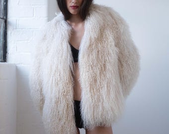 Könnte beinhalten: Ein cremefarbener, zotteliger Kunstfellmantel. Der Mantel hat einen langen, strukturierten Flor und eine lockere Passform. Das Model trägt einen schwarzen BH und einen schwarzen Slip. Der Hintergrund ist eine weiße Wand und ein Fenster.
