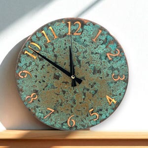 May include: A round wall clock with a textured, teal and copper-colored face. The clock features copper-colored numerals and black hands. The clock is displayed on a wooden surface against a white wall.