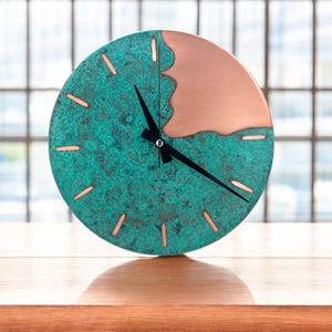 May include: A round clock with a copper and teal face. The clock face has a textured teal finish on the bottom half and a smooth copper finish on the top half. Black clock hands and copper-colored markers indicate the time.