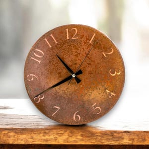May include: A round, copper-colored clock with a distressed finish. The clock face has black hands and white Roman numeral markings. The clock is sitting on a wooden surface.