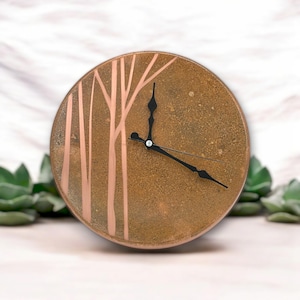 May include: A round, brown, ceramic clock with a copper-colored tree design. The clock has black hands and a simple design.