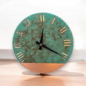 May include: Round wall clock with a teal and copper finish. The clock face has Roman numerals and black hands. The bottom half of the clock is copper-colored, and the top half is teal with a textured appearance.