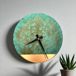 May include: Round wall clock with a distressed turquoise and gold face. The clock has black hands and a gold-colored bottom section. The clock is mounted on a gray wall, with a small potted plant in the lower right corner.