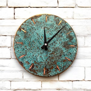 May include: A round, teal-colored wall clock with copper-colored accents. The clock face has a distressed, aged look and features black hands.