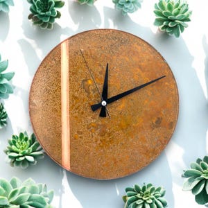 Puede incluir: Un reloj redondo de color oxidado con una franja de color cobre y agujas negras. El reloj está rodeado de pequeñas plantas verdes.