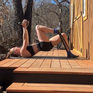 Puede incluir: Una mujer est&aacute; haciendo un puente de gl&uacute;teos con una banda de resistencia y una kettlebell. Lleva un sujetador deportivo negro y pantalones cortos negros. El ejercicio se realiza en una plataforma de madera.