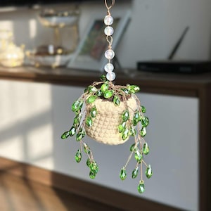 May include: A hanging crochet planter with a beaded chain and green crystal accents. The beige pot holds a faux plant with cascading green crystal leaves. The planter is suspended from a copper-colored wire.