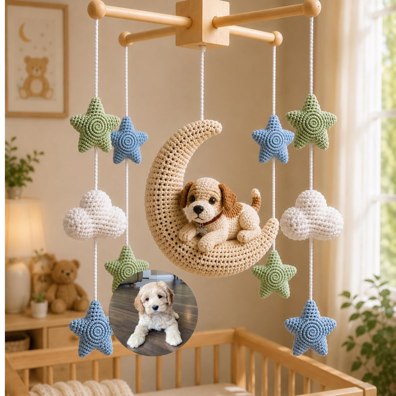 Puede incluir: Un m&oacute;vil colgante para guarder&iacute;a con una media luna beige de ganchillo sobre la que descansa una peque&ntilde;a figura de cachorro marr&oacute;n y blanco. El m&oacute;vil incluye estrellas de ganchillo en verde salvia y azul, y nubes blancas, suspendidas de un marco de madera.