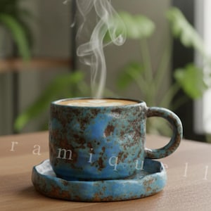 May include: A blue ceramic coffee cup and saucer set with a textured, rustic finish. The cup contains a light-coloured beverage, with steam rising. The set is on a wooden table, with plants in the background. The word "Ceramiquelle" is visible.