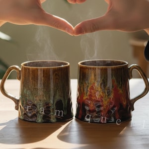 Könnte beinhalten: Zwei Keramikbecher mit brauner und grüner Glasur, die mit heißer Flüssigkeit dampfen. Die Becher stehen auf einem Holztisch, mit Händen, die ein Herz formen. Die Becher tragen das Wort "Ceramique".
