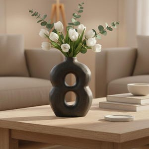 May include: A modern living room scene featuring a wooden coffee table with a dark gray vase in the shape of the number eight. The vase holds white tulips and greenery. A lamp and two beige sofas are in the background.