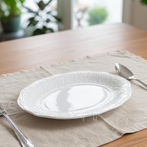 May include: A white oval plate with a decorative embossed pattern, a silver fork, and a spoon are arranged on a beige linen placemat atop a wooden table. The scene is set in a well-lit room with plants visible in the background.