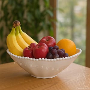Könnte beinhalten: Eine cremefarbene Obstschale aus Keramik, gefüllt mit Bananen, roten Äpfeln, Trauben und einer Orange. Die Schale hat einen Perlenrand und das Wort "Ceramique" ist auf der Seite aufgedruckt.