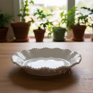 Peut inclure: Un plat de service en céramique blanche avec un bord festonné. Le plat est de forme ronde et peu profond, posé sur une surface en bois. Le mot "quell" est partiellement visible. Des plantes en pot sont visibles en arrière-plan.