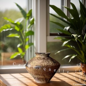 May include: A brown ceramic vase with a textured surface. The vase is sitting on a wooden table in front of a window.