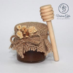 Può includere: Un piccolo barattolo di vetro pieno di un liquido scuro, probabilmente miele, è adornato con un coperchio di tela di juta, spago e fiori secchi. Un cucchiaio di miele di legno è appoggiato accanto al barattolo. Un regalo affascinante.