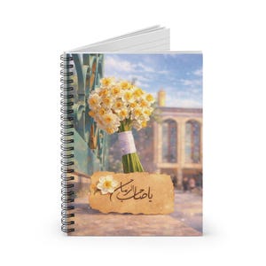 May include: Spiral-bound notebook featuring a bouquet of yellow and white flowers tied with a white ribbon. The cover displays a scenic background with a building and text in Arabic script on a parchment-like label.