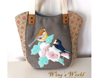 May include: A gray canvas tote bag with a floral design featuring two birds. The bag has brown leather accents and cream-colored straps. The text "Wing's World Dream Work Series" is printed on the bottom of the bag.