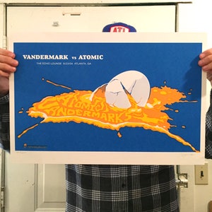 May include: A blue poster with the text "VANDERMARK vs ATOMIC" and "THE ECHO LOUNGE 6/23/04 ATLANTA, GA". The poster features a cracked egg with orange yolk spilling out, and the words "ATOMIC" and "VANDERMARKS" in the yolk.