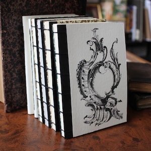 Op de afbeelding: Een stapel kleine, handgemaakte boeken met zwarte ruggen en witte pagina's. De kaft van het voorste boek heeft een zwart-wit sierlijk ontwerp. Op de achtergrond is een groter, antiek boek met een bruine kaft te zien.