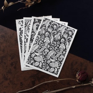 Könnte beinhalten: Ein Stapel schwarz-weiß gemusterter Karten mit floralen und ornamentalen Designs. Die Karten liegen auf einer dunkelbraunen Holzoberfläche, mit dunklem Hintergrund und einer getrockneten Rose.