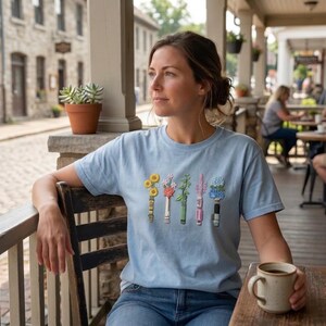 K&ouml;nnte beinhalten: Ein hellblaues Kurzarm-T-Shirt mit einem Grafikdruck von f&uuml;nf vertikalen Gl&uuml;hbirnen, die jeweils ein anderes farbenfrohes Blumenarrangement mit gelben, rosa, gr&uuml;nen, violetten und blauen floralen Elementen auf der Brust zeigen.