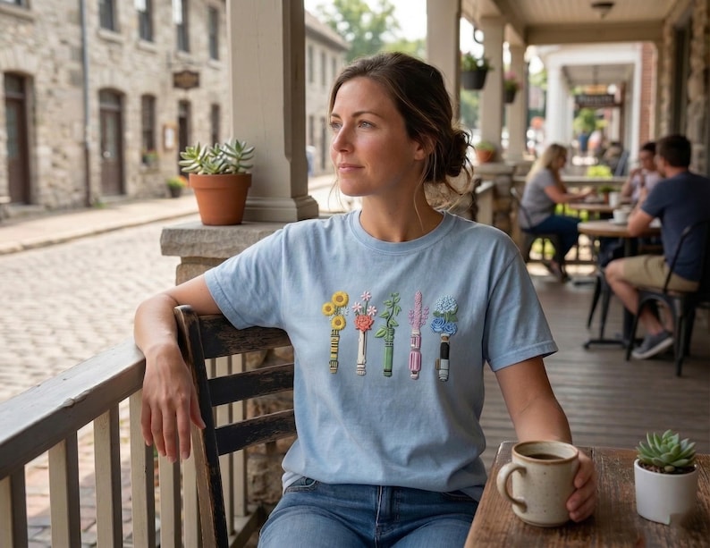 K&ouml;nnte beinhalten: Ein hellblaues Kurzarm-T-Shirt mit einem Grafikdruck von f&uuml;nf vertikalen Gl&uuml;hbirnen, die jeweils ein anderes farbenfrohes Blumenarrangement mit gelben, rosa, gr&uuml;nen, violetten und blauen floralen Elementen auf der Brust zeigen.
