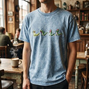 May include: A light blue acid-wash t-shirt featuring a horizontal row of four embroidered green lizard characters in various poses, each accented with small white star details across the centre of the chest.