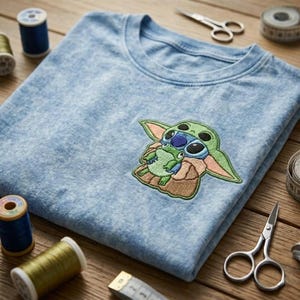 May include: A light blue acid-wash t-shirt featuring an embroidered patch of a green alien character holding a small green frog. The shirt is surrounded by sewing supplies including blue and gold thread spools, metal scissors, and a measuring tape.