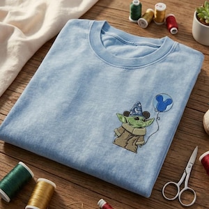 May include: A light blue t-shirt featuring an embroidered graphic of a green creature wearing a blue wizard hat with stars and moons, holding a blue balloon shaped like a mouse head. The embroidery is positioned on the left chest area.