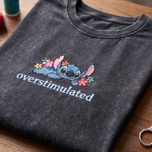K&ouml;nnte beinhalten: Ein anthrazitfarbenes T-Shirt in Acid-Wash-Optik mit einer Stickerei der blauen Figur Stitch, umgeben von rosa, gelben und blauen Blumen. Unter der Figur ist das Wort overstimulated in hellblauen Buchstaben eingestickt.