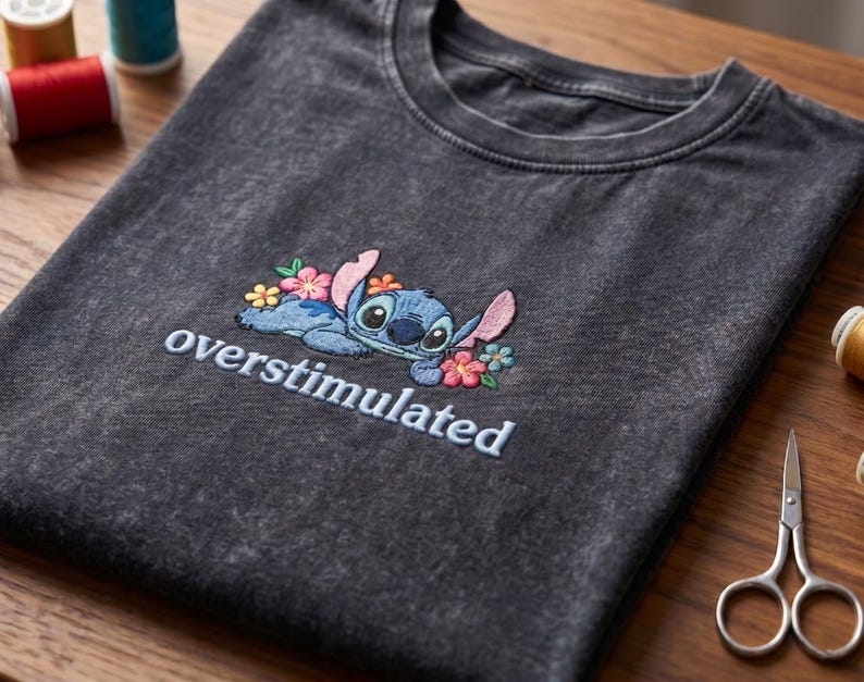 K&ouml;nnte beinhalten: Ein anthrazitfarbenes T-Shirt in Acid-Wash-Optik mit einer Stickerei der blauen Figur Stitch, umgeben von rosa, gelben und blauen Blumen. Unter der Figur ist das Wort overstimulated in hellblauen Buchstaben eingestickt.
