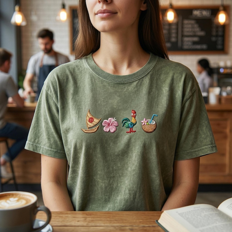 Pode incluir: Uma t-shirt verde-s&aacute;lvia com efeito de lavagem mineral, apresentando uma fila de quatro &iacute;cones bordados: um pequeno veleiro de madeira, uma flor de hibisco rosa, um galo colorido e um coco com uma palhinha azul no centro do peito.