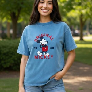 Puede incluir: Una camiseta azul de manga corta con un gr&aacute;fico de Mickey Mouse con pantalones cortos rojos y zapatos blancos. El texto "ORIGINAL" est&aacute; arqueado encima, "19" y "28" a los lados, y "MICKEY" est&aacute; impreso en rojo debajo del gr&aacute;fico.