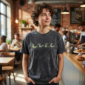 May include: A charcoal grey acid-wash t-shirt featuring a horizontal row of four small, green, cartoon-style lizard illustrations. Each lizard is depicted in a different pose with tiny white star accents scattered around the figures.