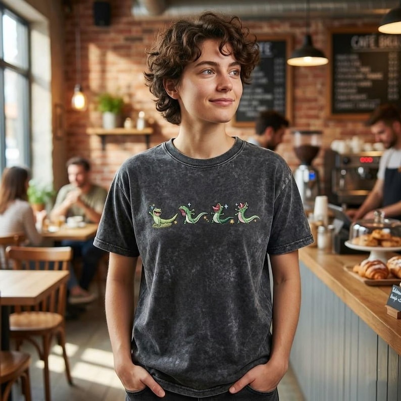 May include: A charcoal grey acid-wash t-shirt featuring a horizontal row of four small, green, cartoon-style lizard illustrations. Each lizard is depicted in a different pose with tiny white star accents scattered around the figures.