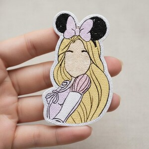 Może przedstawiać: Haftowana naszywka przedstawiająca postać z kresk&oacute;wki z długimi blond włosami, r&oacute;żową kokardą i czarnymi uszami myszy. Postać ma na sobie jasnofioletową sukienkę i trzyma r&oacute;żowy pędzel. Naszywka ma białą obw&oacute;dkę.