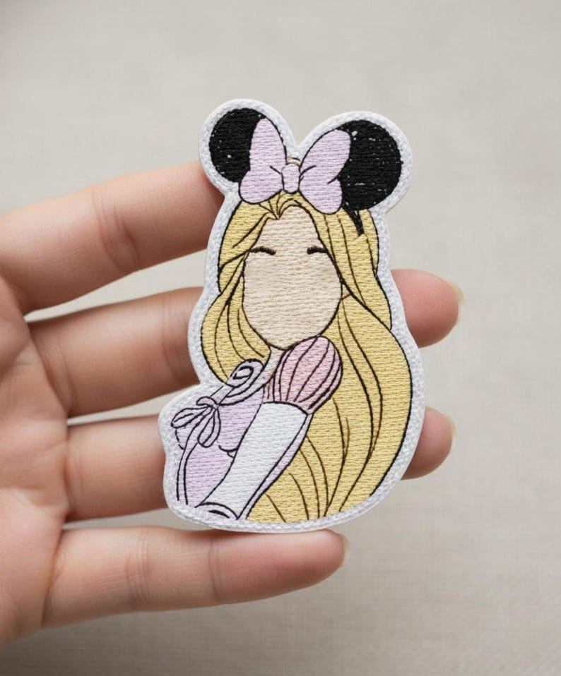 Może przedstawiać: Haftowana naszywka przedstawiająca postać z kresk&oacute;wki z długimi blond włosami, r&oacute;żową kokardą i czarnymi uszami myszy. Postać ma na sobie jasnofioletową sukienkę i trzyma r&oacute;żowy pędzel. Naszywka ma białą obw&oacute;dkę.