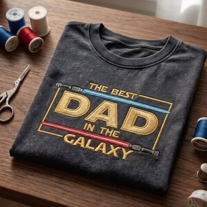 May include: A charcoal gray t-shirt featuring an embroidered design with the text THE BEST DAD IN THE GALAXY in gold letters. The text is framed by a blue light saber at the top and a red light saber at the bottom, creating a rectangular border.