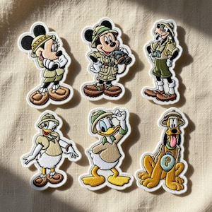 Op de afbeelding: Set van zes geborduurde patches met Disney-personages in safari-outfits. Inclusief Mickey en Minnie Mouse, Goofy, Donald Duck en Pluto. Elke patch heeft een witte rand en is ontworpen met levendige kleuren en gedetailleerde stiksels.