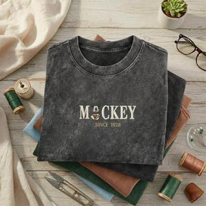 May include: A charcoal gray crewneck t-shirt with a vintage-style wash. The center of the chest features embroidered cream-colored text that reads MOCKEY with a small cartoon mouse icon replacing the letter O, followed by the text SINCE 1928.