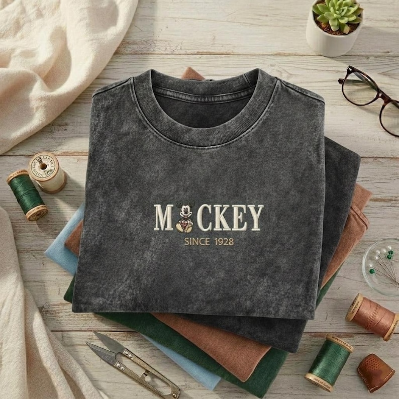 May include: A charcoal gray crewneck t-shirt with a vintage-style wash. The center of the chest features embroidered cream-colored text that reads MOCKEY with a small cartoon mouse icon replacing the letter O, followed by the text SINCE 1928.