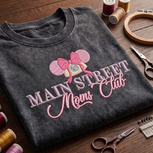 Może przedstawiać: Ciemnoszary t-shirt z efektem sprania, ozdobiony haftem przedstawiającym telefon w stylu vintage z r&oacute;żowymi uszami myszy i kokardą. Napis MAIN STREET Moms Club wykonano jasnofioletową i r&oacute;żową nicią na środku klatki piersiowej.