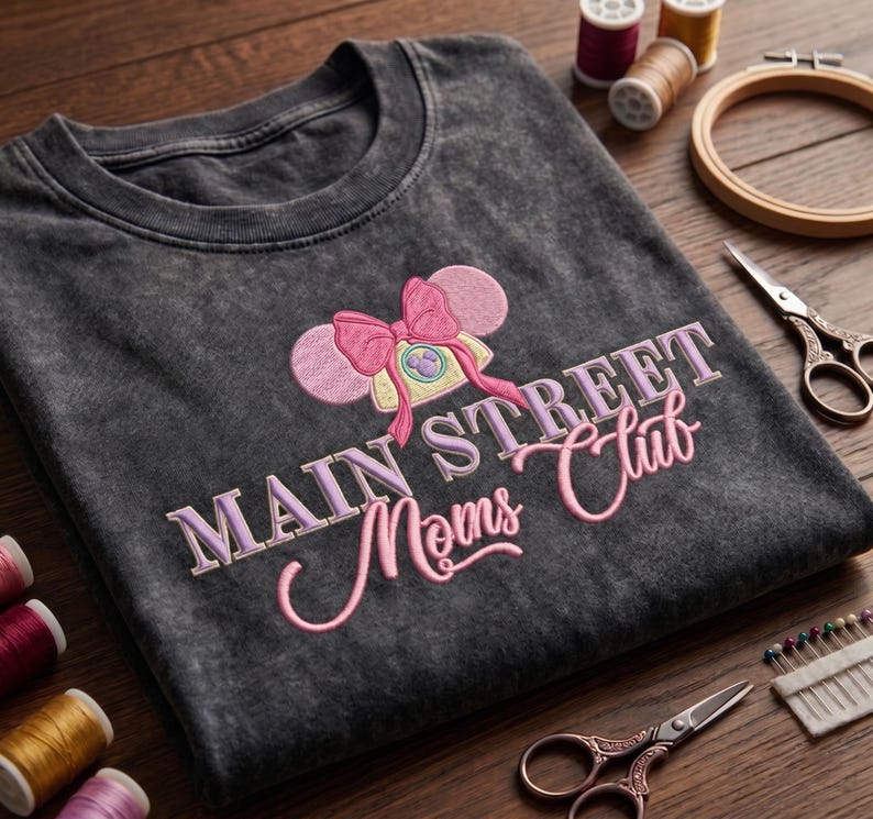 Może przedstawiać: Ciemnoszary t-shirt z efektem sprania, ozdobiony haftem przedstawiającym telefon w stylu vintage z r&oacute;żowymi uszami myszy i kokardą. Napis MAIN STREET Moms Club wykonano jasnofioletową i r&oacute;żową nicią na środku klatki piersiowej.