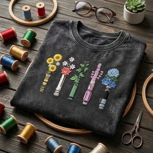 K&ouml;nnte beinhalten: Ein anthrazitfarbenes T-Shirt in Acid-Wash-Optik mit f&uuml;nf Stickereien, die Lichtschwertgriffe zeigen, welche mit verschiedenen Blumen wie Sonnenblumen, einer Rose, gr&uuml;nem Blattwerk, Lavendel und blauen Hortensien verziert sind.